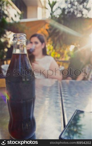 Glass bottle of cold cola soda on metal table. A woman drinking in the background. Sun Shining.