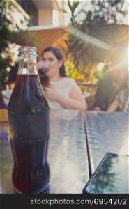 Glass bottle of cold cola soda on metal table. A woman drinking in the background. Sun Shining.