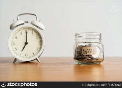 Glass bottle which has coin and a white retro alarm clock on a brown wooden table. Investment business, retirement, finance and saving money for future concept.