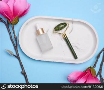 Glass bottle with a pipette, a stone face massager on a marble white stand and a branch with flowers, top view
