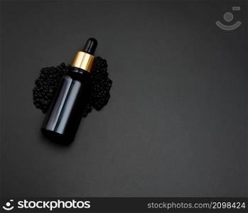 glass bottle with a pipette lies on a heap of black caviar, black background. Natural cometic. Product branding, top view