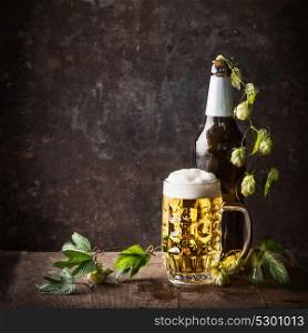 Glass bottles and mug of beer with cap of foam and hops on table at dark rustic background, front view, Still life , close up