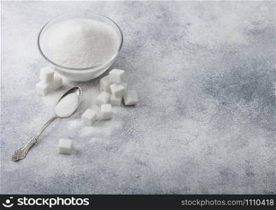 Glass bowl of natural white refined sugar with cubes and silver spoon on light background.