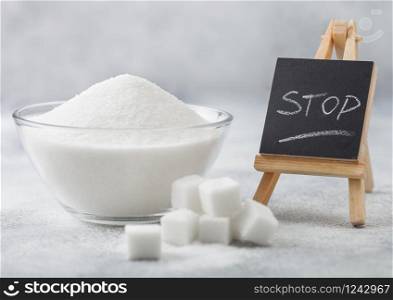 Glass bowl of natural white refined sugar with cubes with silver spoon on light background with STOp letters on mini chalkboard. Unhealthy food concept. Macro