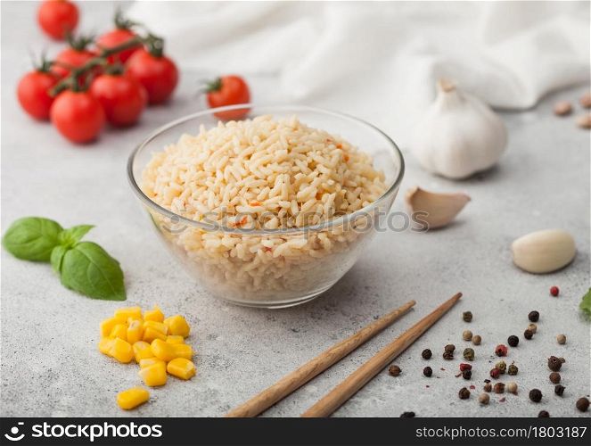 Glass bowl with boiled long grain basmati rice with vegetables on light background with sticks and tomatoes with corn, garlic and basil.