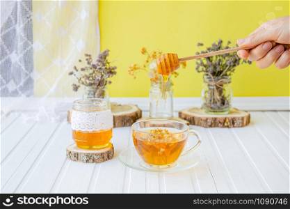 Glass cup of tea with linden in natural organic herbs and a jar of honey on a white wooden table. Increase immunity in the cool season.. Glass cup of tea with linden in natural organic herbs and a jar of honey on a white wooden table.