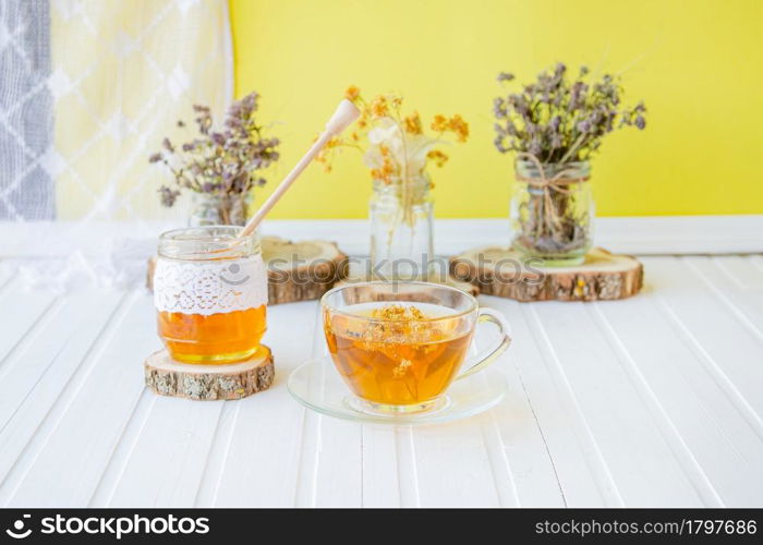 Glass cup of tea with linden in natural organic herbs and a jar of honey on a white wooden table. Increase immunity in the cool season.. Glass cup of tea with linden in natural organic herbs and a jar of honey on a white wooden table.