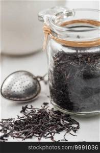 Glass jar of black loose organic tea with ceramic teapot and vintage strainer infuser on light background.