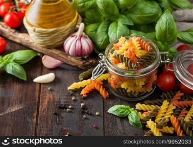 Glass jar of fresh raw tricolor fusilli pasta with oil and garlic, basil plant and tomatoes on wooden background. Top view