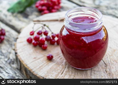 Glass jar of homemade viburnum jam with fresh viburnum berries on a wooden table. Source of natural vitamins. Used in folk medicine. Autumn harvest.. Glass jar of homemade viburnum jam with fresh viburnum berries on a wooden table. Source of natural vitamins. Used in folk medicine.