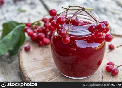 Glass jar of homemade viburnum jam with fresh viburnum berries on a wooden table. Source of natural vitamins. Used in folk medicine. Autumn harvest.. Glass jar of homemade viburnum jam with fresh viburnum berries on a wooden table. Source of natural vitamins. Used in folk medicine.
