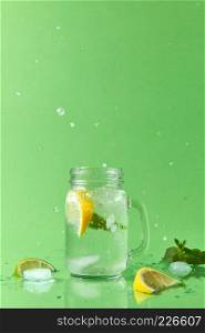 Glass jar on a green table with homemade sparkling lemonade with slices of lime and lemon, green leaves of mint. Splash lemonade from a glass and slices lime, lemon on a green table .. Splash lemonade in different directions from a mason jar with natural lemonade handmade. Drops of liquid and pieces of lemon and lime on a green table with place for text.