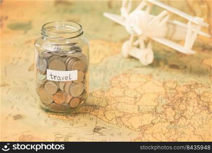 Glass jar with coins and travel inscription on world map with wooden toy plane on table. Travel concept and saving money.