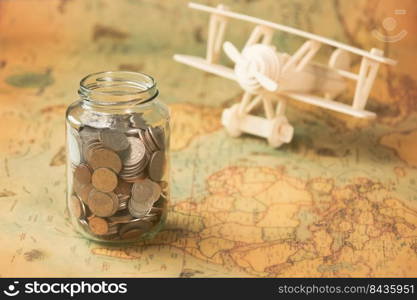 Glass jar with coins on world map with wooden toy plane on table. Travel concept and saving money.