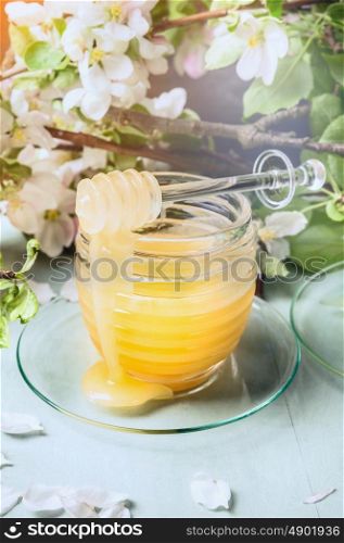 Glass jar with honey and dipper over fresh garden blossom on light blue wooden background