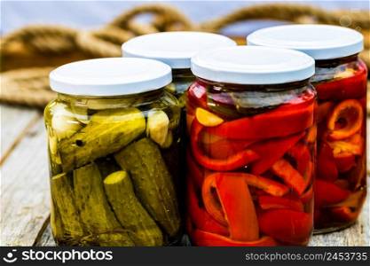 Glass jars with pickled red bell peppers and pickled cucumbers (pickles) isolated. Jars with variety of pickled vegetables. Preserved food concept in a rustic composition.