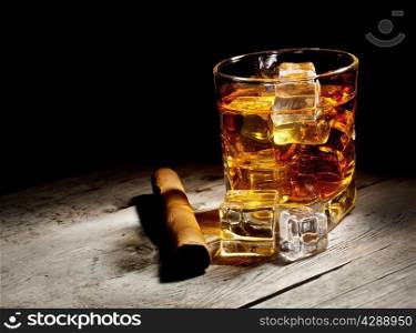 Glass of aged whiskey with cigar and ice cubes on wooden table