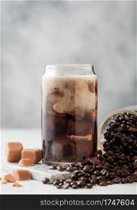 Glass of black iced coffee with fresh milk and jar of raw coffee beans and salted caramel on light table background.