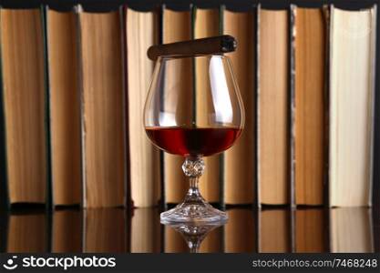 Glass of brandy on a reflective surface with books and cigar