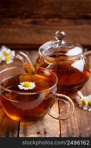 Glass of chamomile tea with full hot teapot and fresh chamomile flowers on vintage old wooden table background