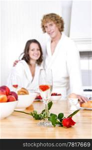 Glass of champagne with a strawberry floating on it, and a red rose on a breakfast counter with a young couple in the background.
