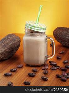 glass of cocoa drink with a green straw, acompanied by dried cacao fruits an cacao beans before an orange background.