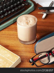 Glass of cocoa on a wooden office table, a book a writing machine and a pair of glasses.