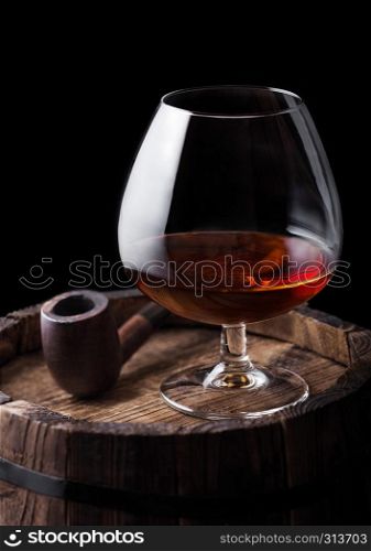 Glass of cognac brandy drink and vintage smoking pipe on top of wooden barrel on black background.