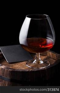 Glass of cognac brandy drink with black coaster on top of wooden barrel on black background.