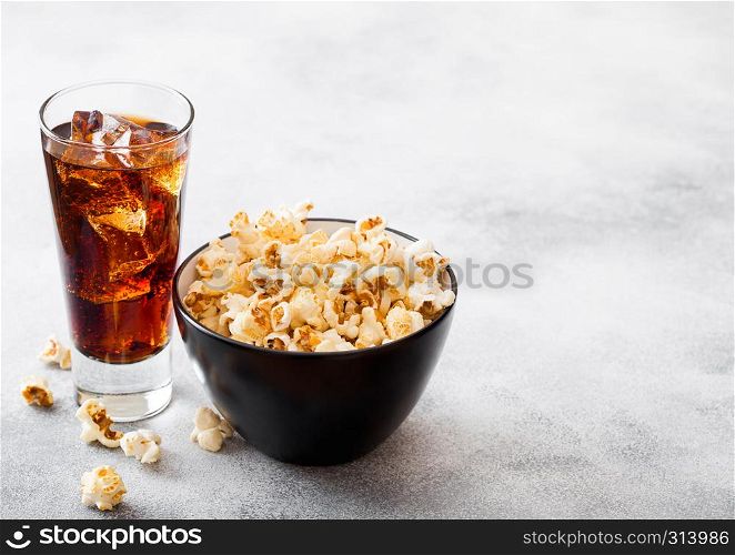 Glass of cola soda drink with ice cubes and black bowl of popcorn snack on stone kitchen background.