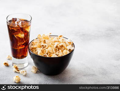 Glass of cola soda drink with ice cubes and black bowl of popcorn snack on stone kitchen background.