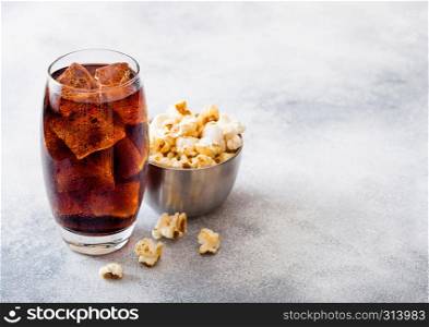 Glass of cola soda drink with ice cubes and steel bowl of popcorn snack on stone kitchen background.