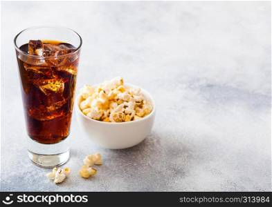 Glass of cola soda drink with ice cubes and whitel bowl of popcorn snack on stone kitchen background.