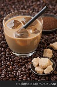 Glass of cold coffee with ice cubes and straw on fresh raw coffee beans background with cane sugar and powder.