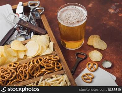 Glass of craft lager beer and opener with box of snacks on brown kitchen table background. Pretzel and crisps and salty potato sticks in vintage wooden box with openers and beer mats.