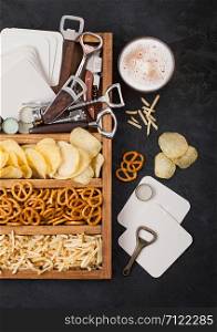 Glass of craft lager beer and opener with box of snacks on black kitchen table background. Pretzel and crisps and salty potato sticks in vintage wooden box with openers and beer mats.