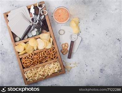 Glass of craft lager beer and opener with box of snacks on light kitchen table background. Pretzel and crisps and salty potato sticks in vintage wooden box with openers and beer mats.