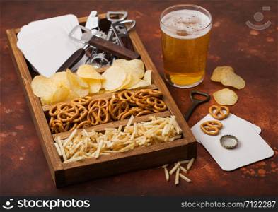Glass of craft lager beer and opener with box of snacks on brown kitchen table background. Pretzel and crisps and salty potato sticks in vintage wooden box with openers and beer mats.