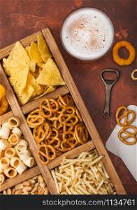 Glass of craft lager beer and opener with box of snacks on brown kitchen background. Pretzel,salty potato sticks, peanuts, onion rings with nachos in vintage box with openers and beer mats.