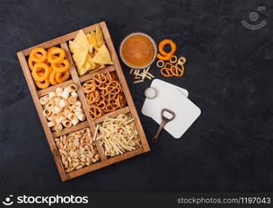 Glass of craft lager beer and opener with box of snacks on dark background. Pretzel,salty potato sticks, peanuts, onion rings with nachos in vintage box with openers and beer mats. Top view