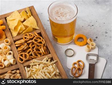 Glass of craft lager beer and opener with box of snacks on light kitchen background. Pretzel,salty potato sticks, peanuts, onion rings with nachos in vintage box with openers and beer mats.