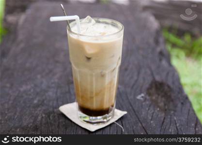 glass of iced coffee laid on piece of wood in the garden.