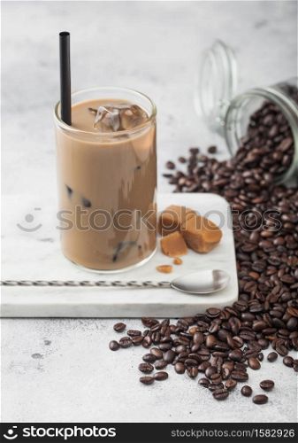 Glass of iced coffee with milk on marble board with jar of coffee beans and salted caramel and long spoon on light table background.