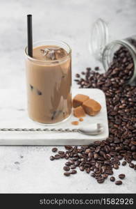 Glass of iced coffee with milk on marble board with jar of coffee beans and salted caramel and long spoon on light table background.