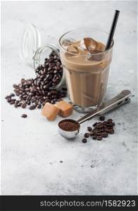 Glass of iced coffee with milk with jar of coffee beans and salted caramel and silver scoop on light table background.