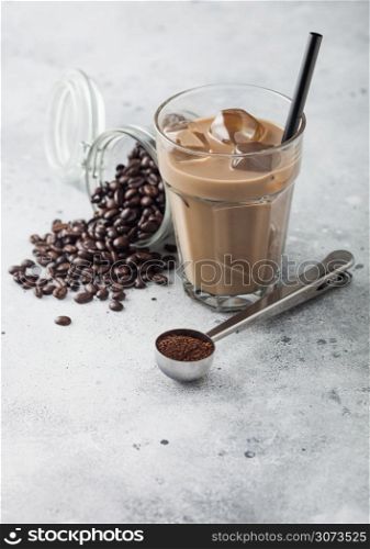 Glass of iced coffee with milk with jar of coffee beans and silver scoop on light table background.