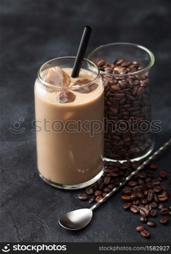 Glass of iced cold coffee and milk with free raw coffee beans in glass container with long spoon on black background.