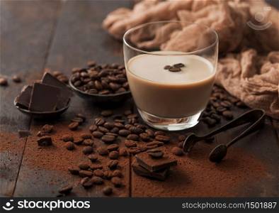 Glass of Irish cream baileys liqueur with ice cubes,coffee beans and powder with dark chocolate and brown cloth on dark wood background. Space for text