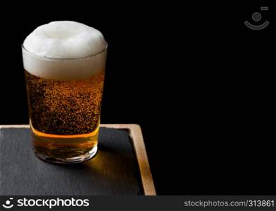 Glass of lager beer with foam and bubbles on stone board on black. Space for text