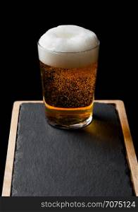 Glass of lager beer with foam and bubbles on stone board on black.